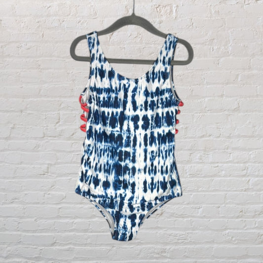 Blue and white tie-dye one-piece swimsuit with cross back detail, displayed on a hanger against a brick wall.