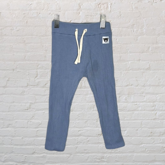 Children's blue ribbed jogger pants with a drawstring and bear patch, hanging against a white brick wall.