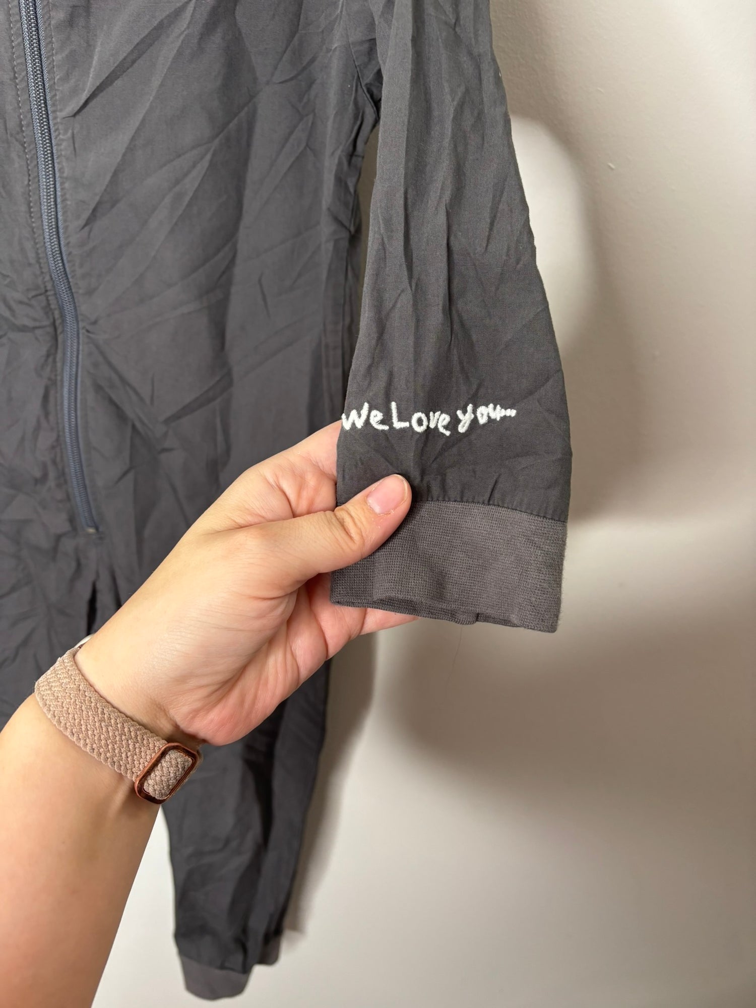 Dark grey coveralls with 'Hotel Cleaning Crew' printed on the back, displayed on a hanger against a white brick wall.