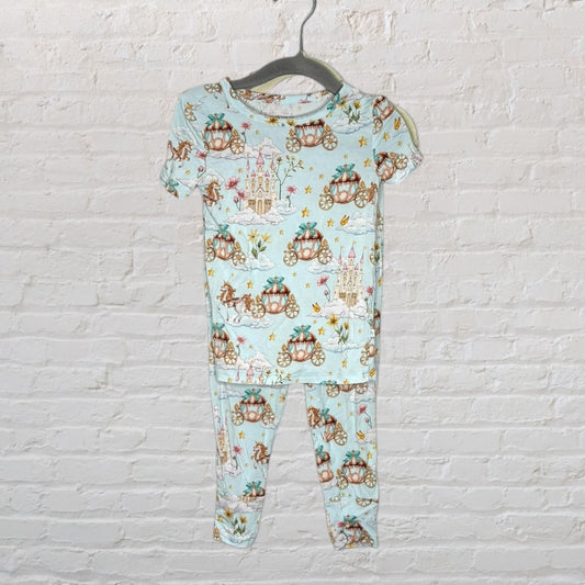 Light blue children's pajama set with fairytale carriage and castle print, hanging on a hanger against a white brick wall.