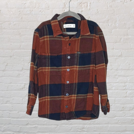 Rust and navy plaid button-up flannel shirt for kids by Abercrombie Kids, size 11/12, displayed on a hanger against a white brick wall.