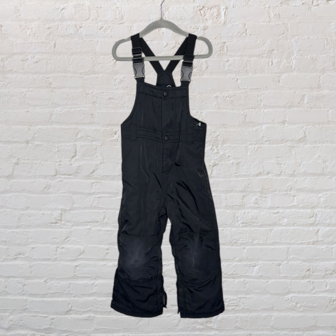 Black children's snow bib overalls with adjustable straps, hanging on a hanger against a white brick wall.