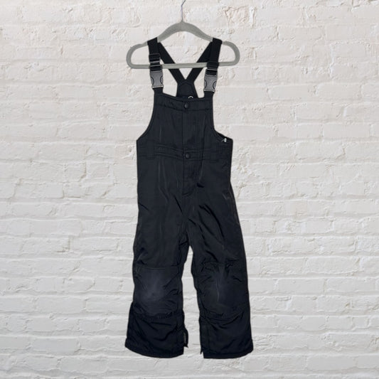 Black children's snow bib overalls with adjustable straps, hanging on a hanger against a white brick wall.