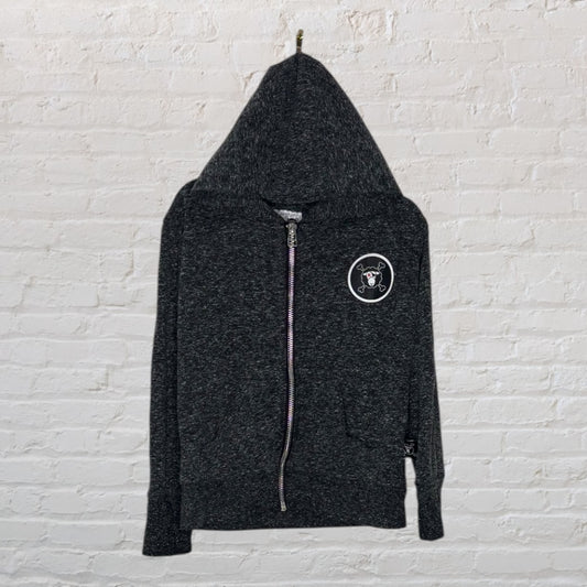 Charcoal zip-up hoodie with a skull and crossbones patch on the left chest, displayed against a white brick wall.