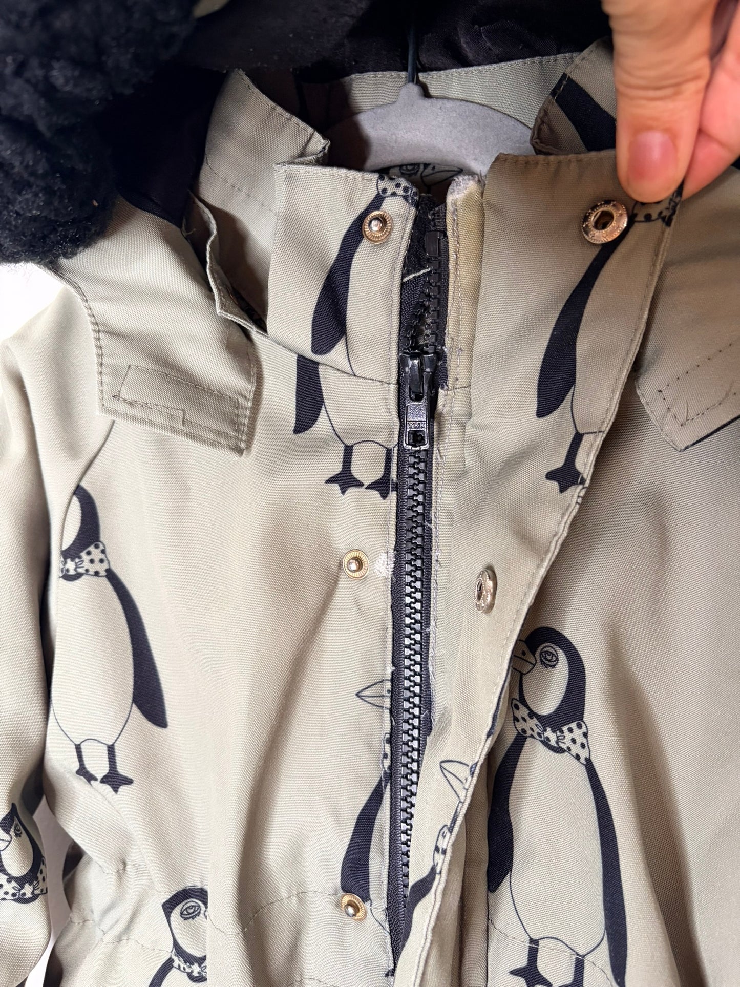 Green hooded parka with black penguin print and faux fur trim, showcased against a white brick wall.