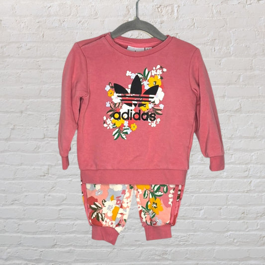 Pink Adidas sweatshirt and pants set with floral design and logo, displayed on a hanger against a white brick wall.