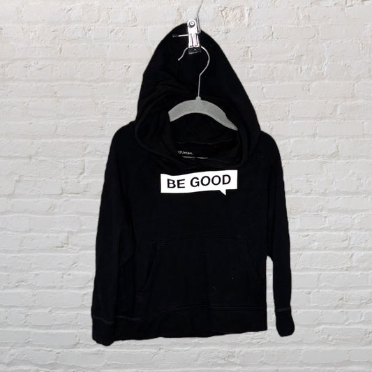 Black hoodie with 'Be Good' graphic on the front, displayed on a hanger against a white brick wall.
