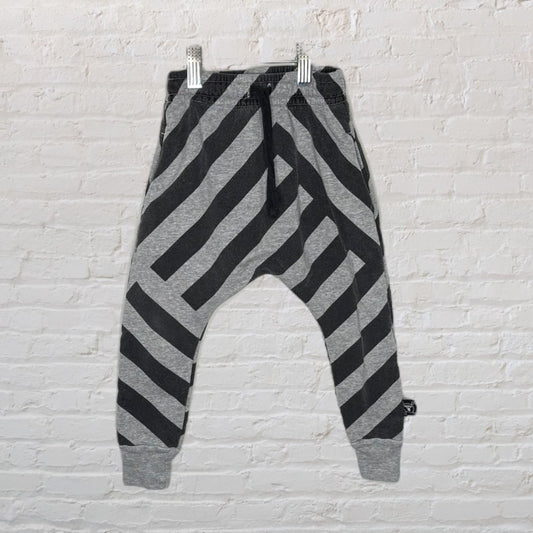 Black and grey diagonal striped harem pants hanging on a hanger against a white brick wall.