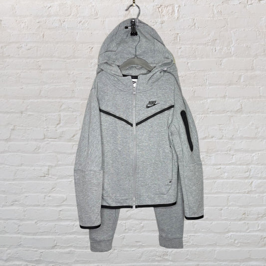 Grey Nike Tech Fleece full zip hoodie and pants set with black trim accents displayed against a white brick wall.