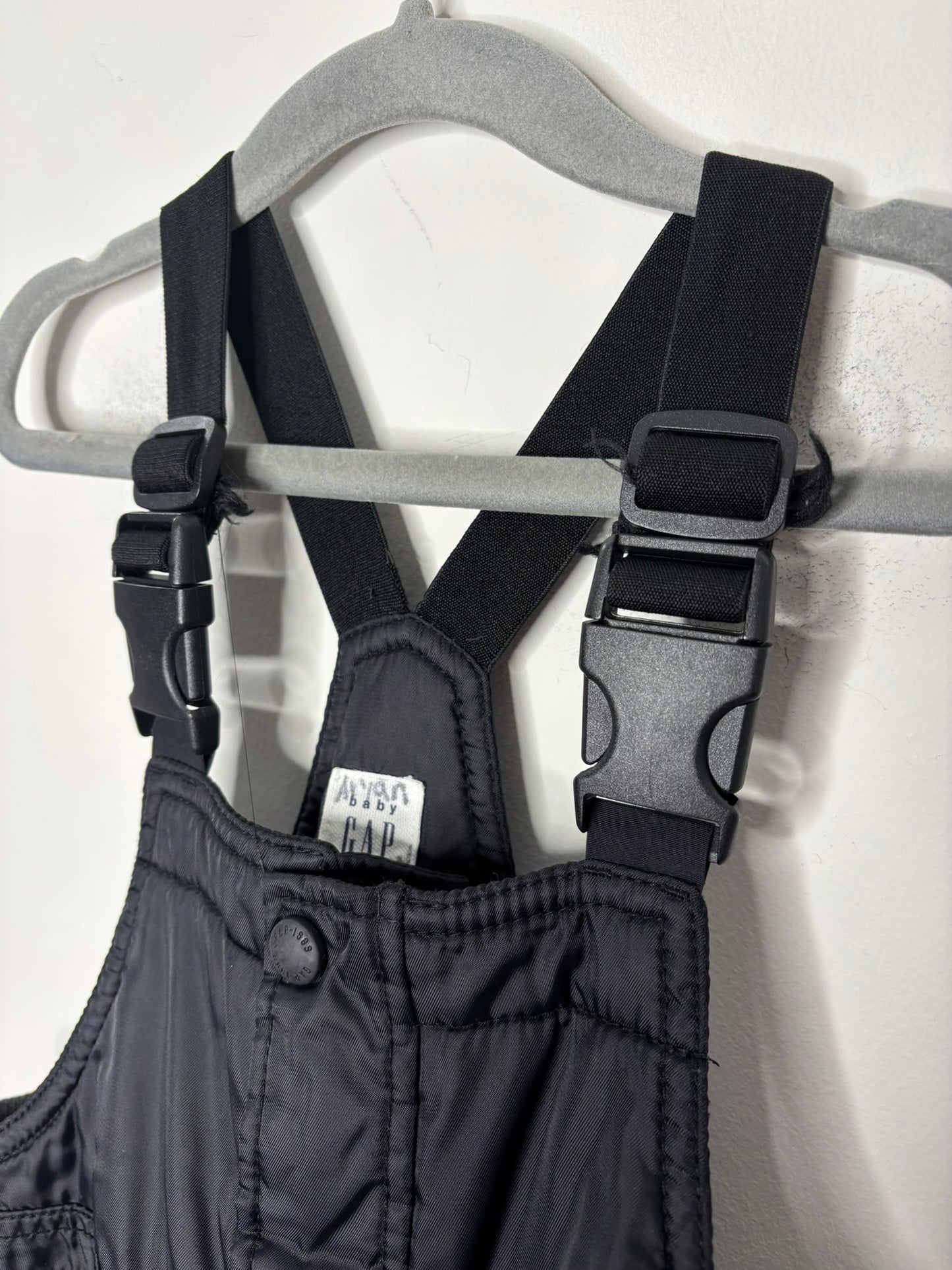 Children's black snow bib overalls with adjustable straps hanging on a hanger.