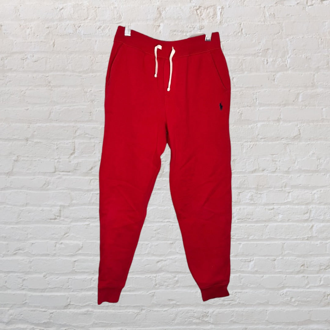 Red drawstring jogger sweatpants with elastic cuffs hanging against a white brick wall.