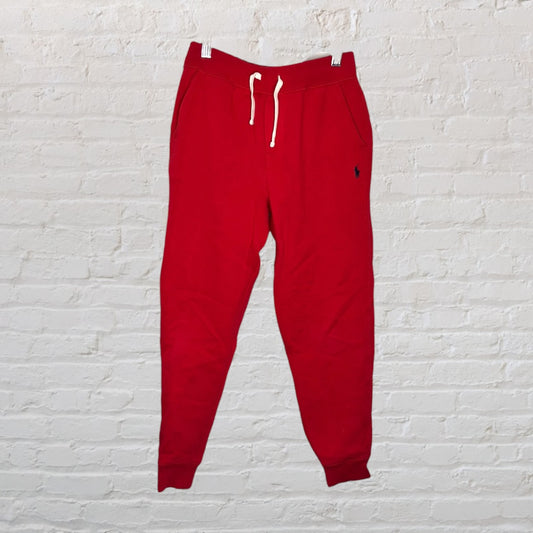 Red drawstring jogger sweatpants with elastic cuffs hanging against a white brick wall.