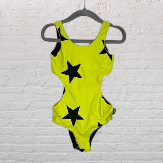 Neon yellow one-piece swimsuit with black star prints, hanging on a hanger against a white brick wall.