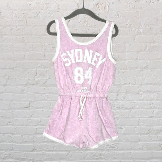 Light pink sleeveless romper with white trim and 'Sydney 84' graphic text, hanging on a black hanger.