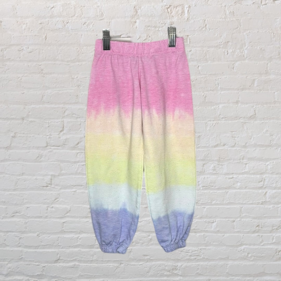 Colorful tie-dye sweatpants with pink, yellow, and blue gradient, on a white brick background.