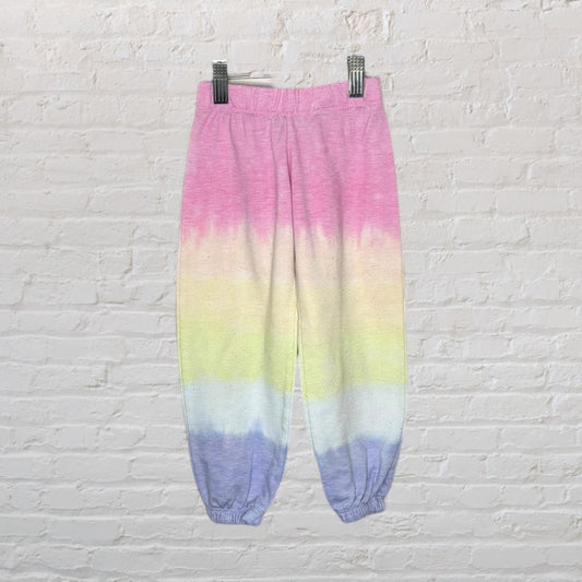 Colorful tie-dye sweatpants with pink, yellow, and blue gradient, on a white brick background.