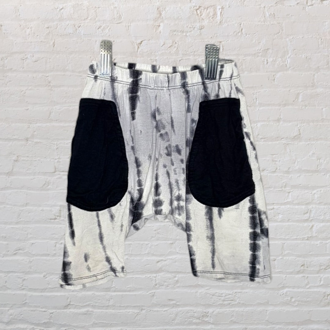 Unisex black and white tie-dye shorts with black pockets, hanging on clips against a white brick wall.
