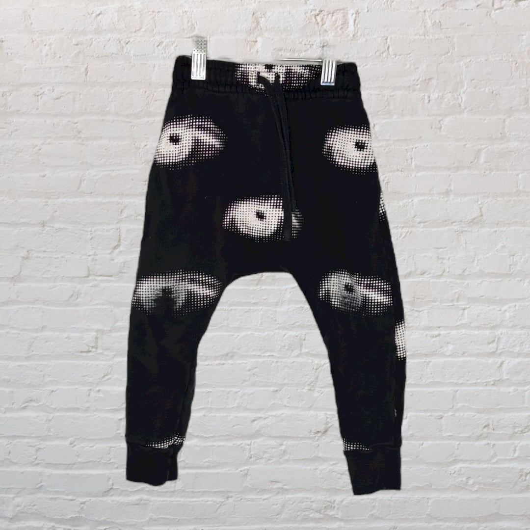 Black jogger pants featuring a white eye print design hanging on a white brick background.