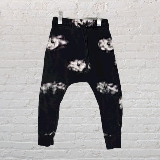 Black jogger pants featuring a white eye print design hanging on a white brick background.