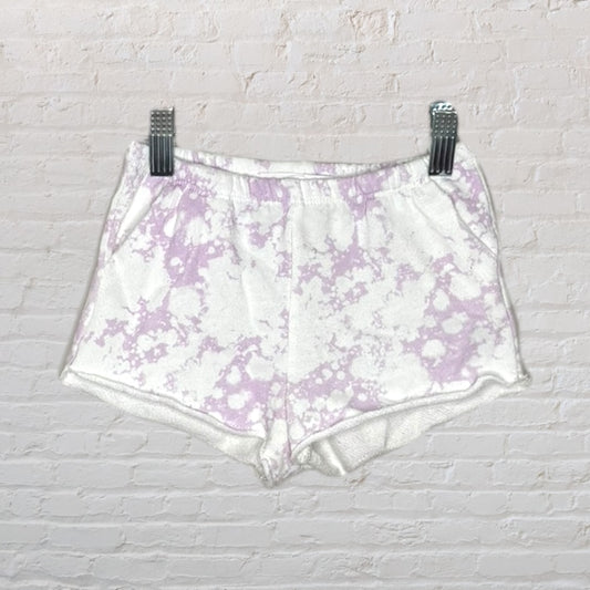 White and lavender tie-dye shorts with elastic waistband, hanging on a clip hanger against a brick wall.