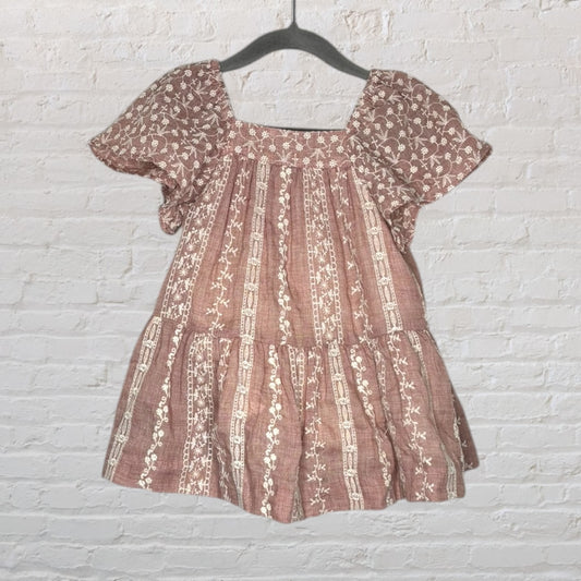 Dusty rose dress with white floral embroidery and puff sleeves, hanging on a dark hanger against a white brick wall.