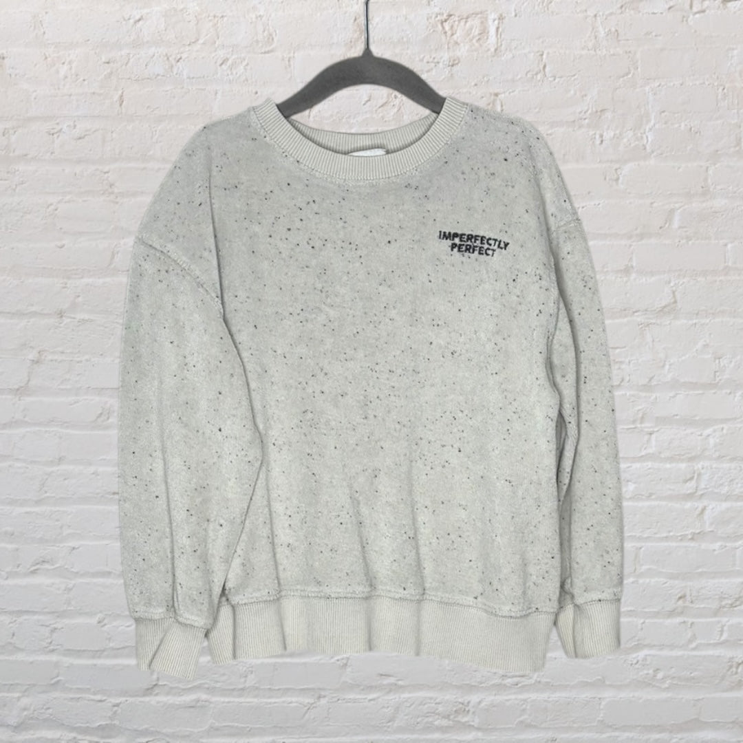 Light grey speckled sweatshirt with 'Imperfectly Perfect' text on the chest, displayed on a hanger against a brick wall.