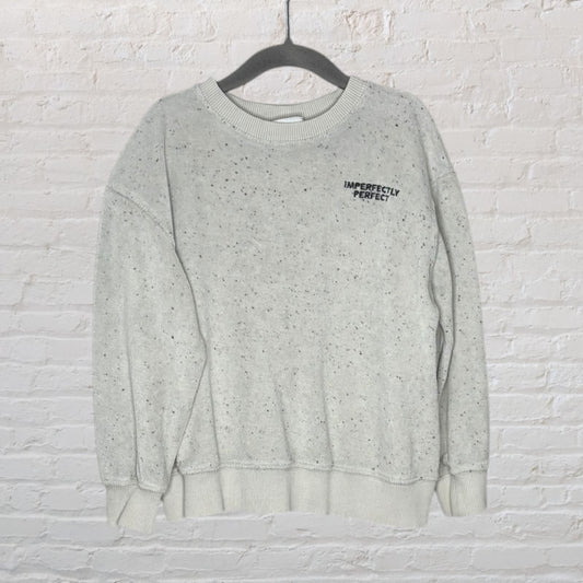 Light grey speckled sweatshirt with 'Imperfectly Perfect' text on the chest, displayed on a hanger against a brick wall.