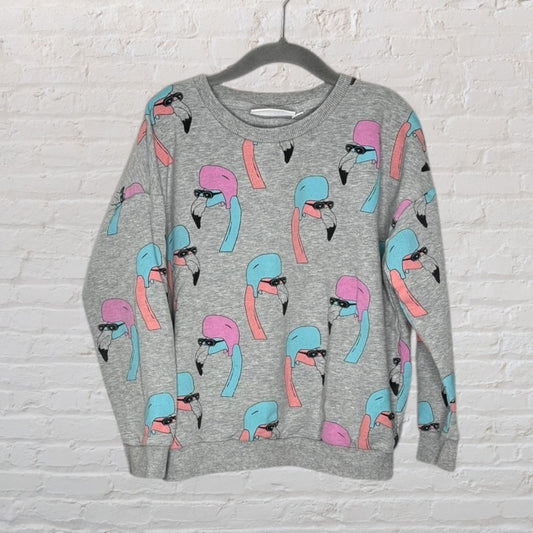 Grey sweatshirt with a colorful flamingo print in pink and blue, hanging on a hanger against a white brick wall.