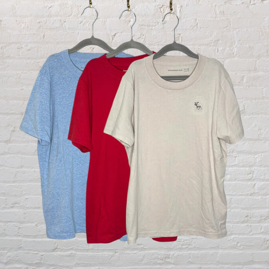 Set of three Abercrombie Kids T-shirts in beige, red, and blue, each hanging on a separate hanger against a white brick wall.