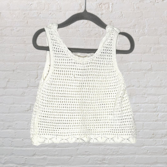 Cream sleeveless crochet knit top hanging on a black hanger against a white brick wall.