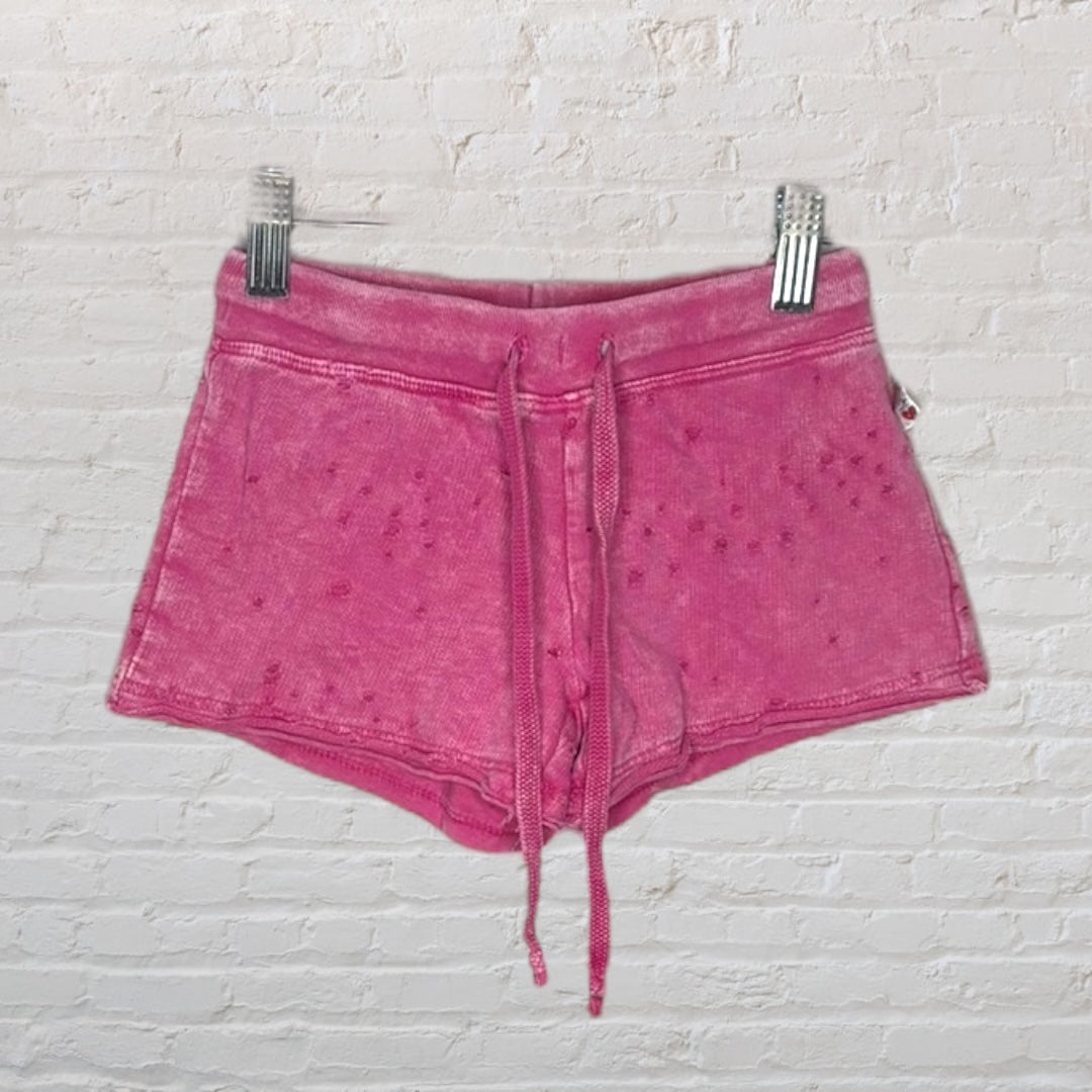 Pink distressed drawstring shorts with a worn-in look on a hanger against a white brick wall.