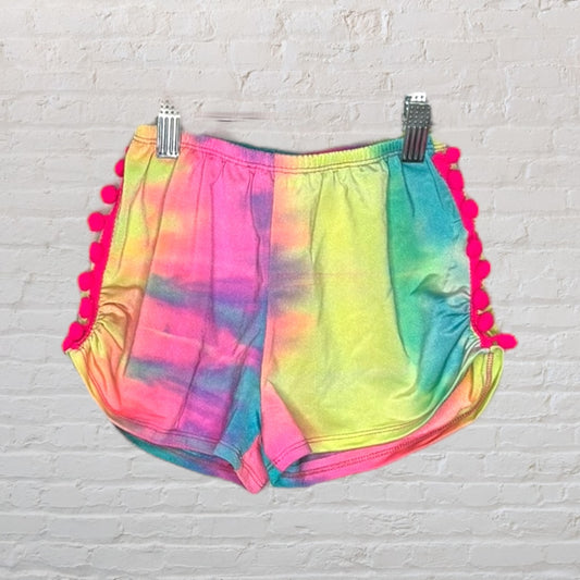 Rainbow tie-dye shorts with pink pom-pom trim along the sides, clipped on a hanger.