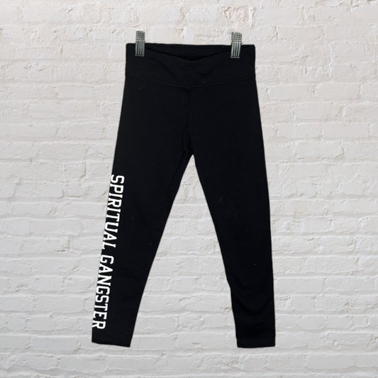 Black Spiritual Gangster leggings with white text on the leg, hanging against a white brick wall.