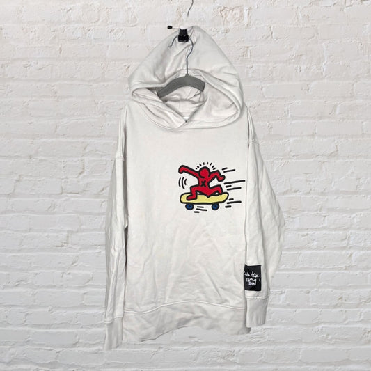 White hoodie with a Keith Haring style red figure on a skateboard graphic, hanging on a brick wall background.
