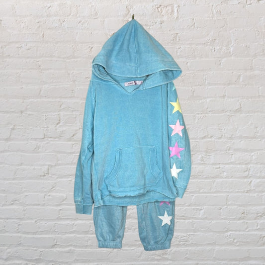 Blue hoodie and joggers set with colorful stars along one sleeve and leg, hanging against a white brick wall.