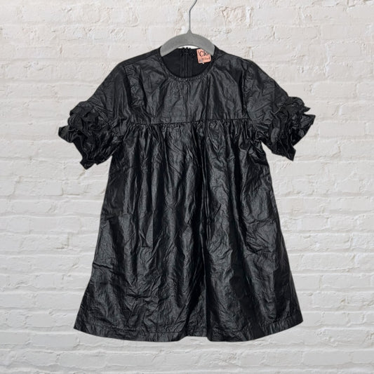 Black short-sleeve dress with ruffled accents and a flowy design, hanging on a hanger against a white brick background.