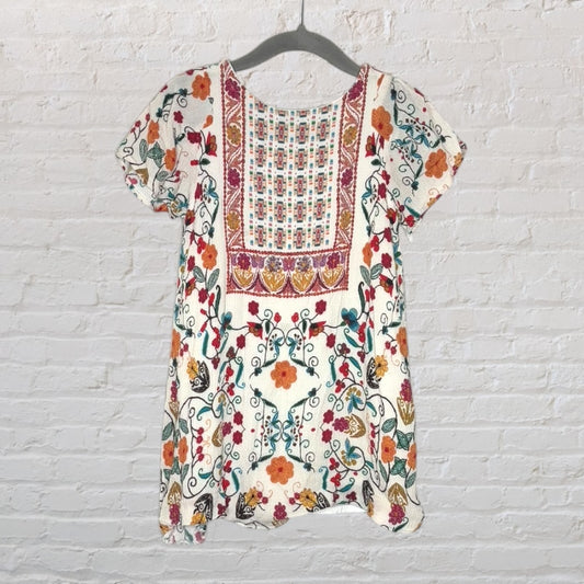 Cream blouse with multicolored floral embroidery and short sleeves hanging on a hanger against a white brick wall.
