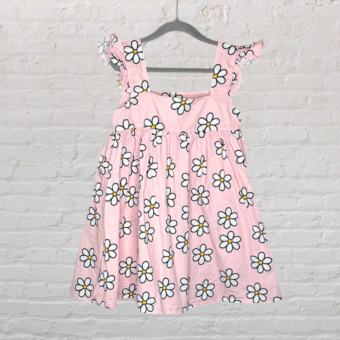 Pink sundress with white daisy print, featuring ruffled straps, hanging on a hanger against a white brick wall.