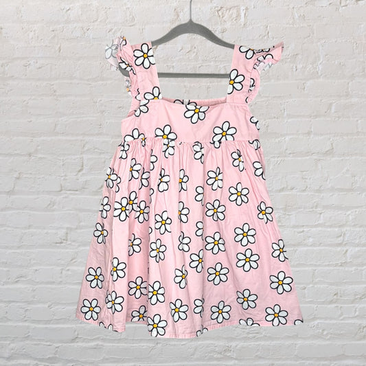 Pink sundress with white daisy print, featuring ruffled straps, hanging on a hanger against a white brick wall.