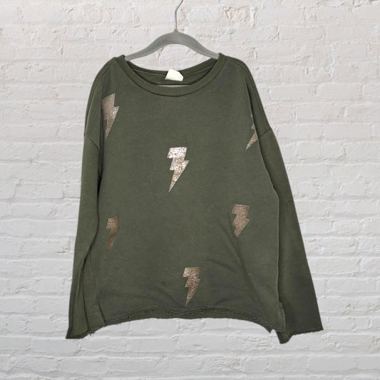 Olive green sweatshirt with silver lightning bolt patterns, hanging on a hanger against a brick wall.