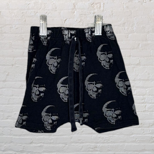 Black knit shorts with grey skull print and drawstring, clipped on a hanger against a brick wall.