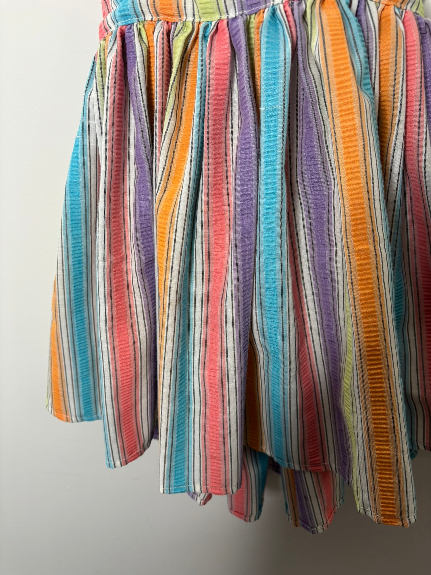A vibrant rainbow-striped sundress with spaghetti straps, hanging on a black hanger against a white brick wall.