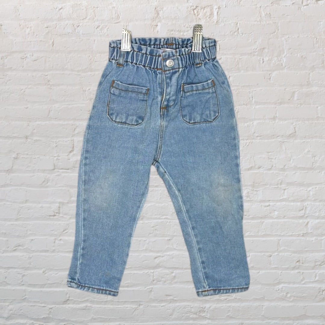 Light blue children's denim jeans with elastic waistband and front pockets, displayed against a white brick wall.