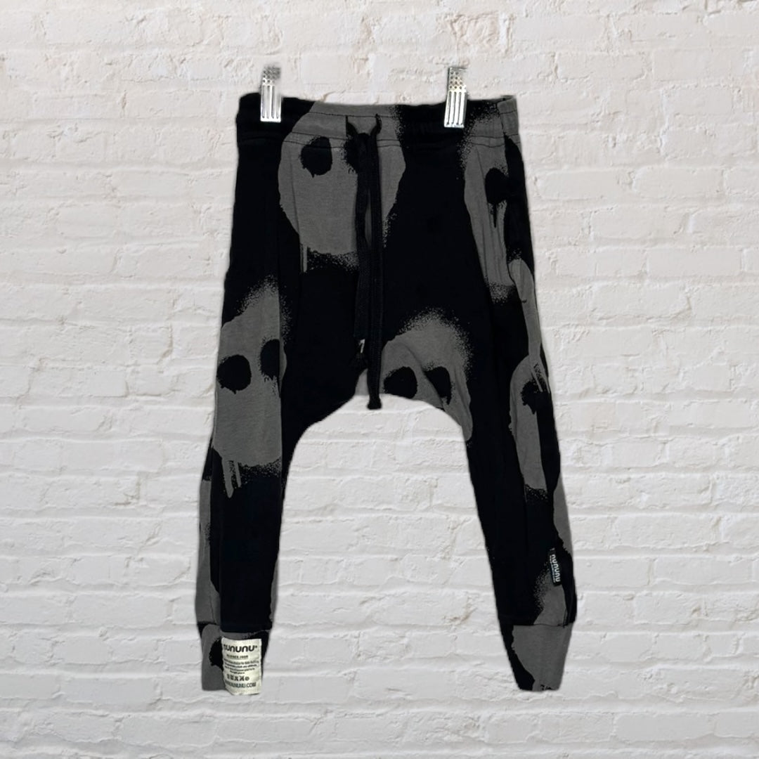 Black and gray drop crotch pants with skull print pattern and drawstring waist by NUNUNU, hanging on a white brick wall.