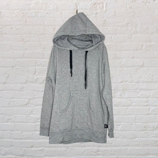 A gray oversized hoodie with black drawstrings and a kangaroo pocket, displayed on a white brick wall.
