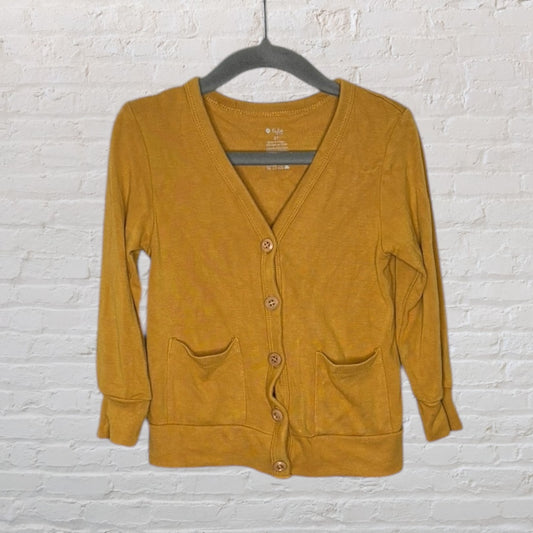 Mustard yellow button-up cardigan with pockets, displayed on a hanger against a brick wall.
