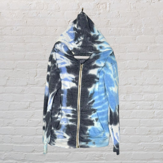 Tie-dye zip-up hoodie in shades of blue, black, and white, displayed on a hanger against a brick wall.