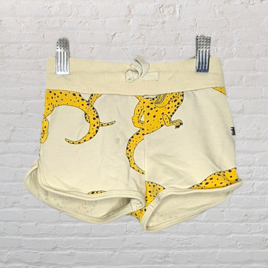 Beige shorts with yellow leopard print design, displayed on clips against a white brick background.