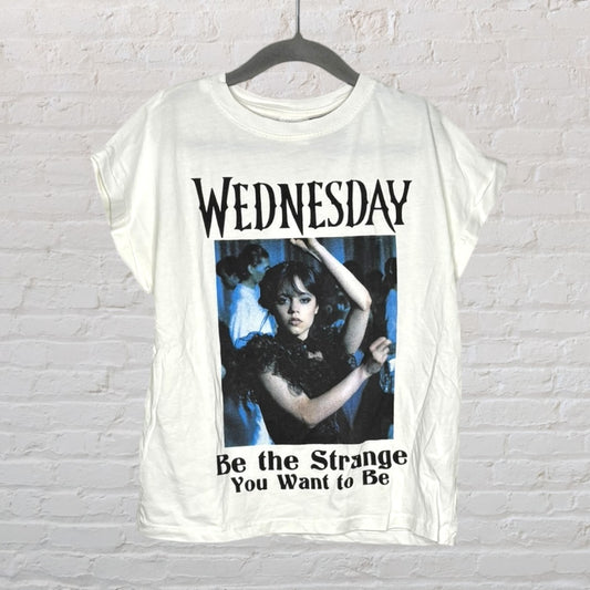 White T-shirt with 'Wednesday' graphic and quote 'Be the Strange You Want to Be' displayed on a hanger.