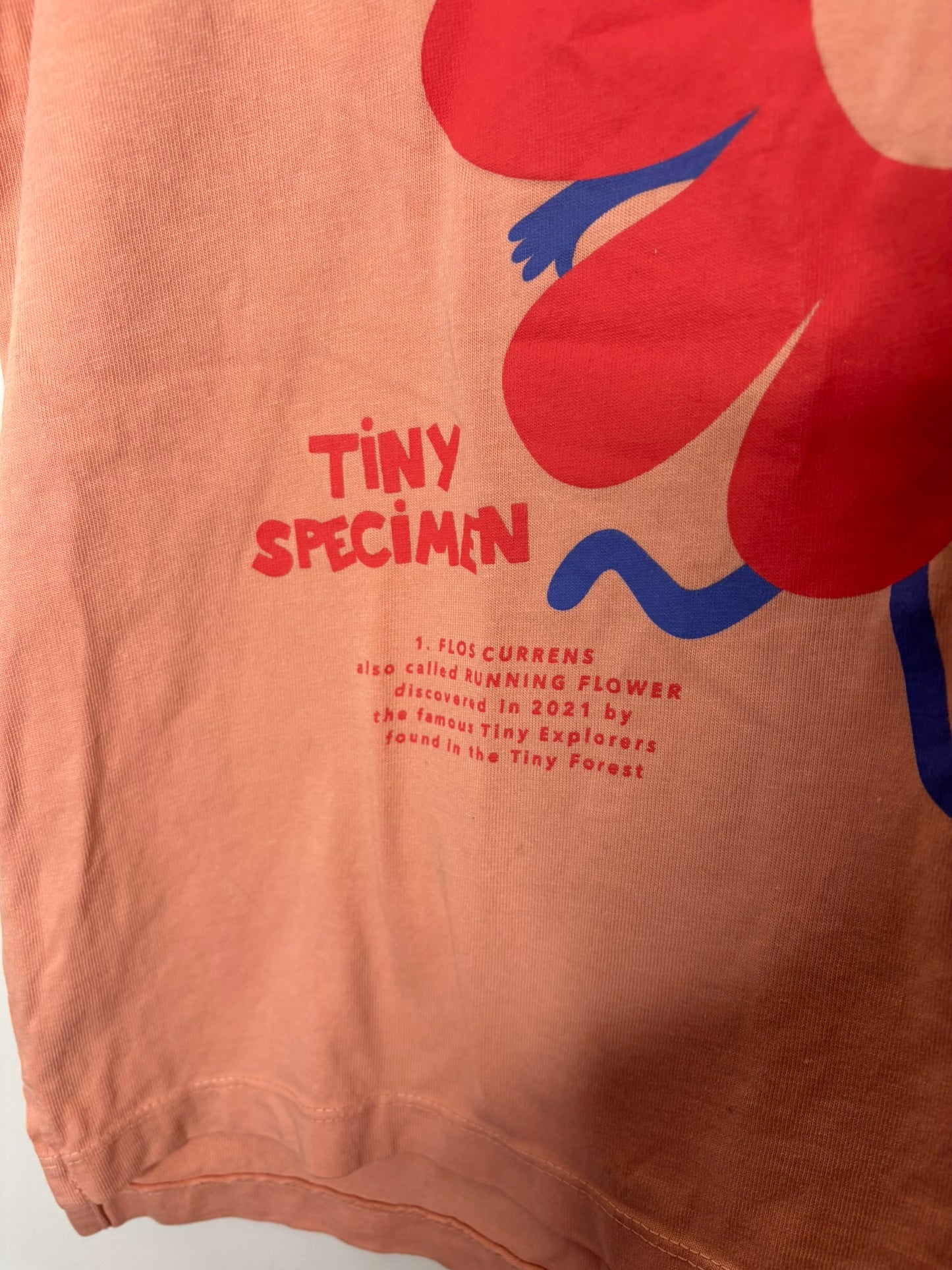 Peach T-shirt featuring a red flower with a smiling face and 'Tiny Specimen' text, hanging on a black hanger.
